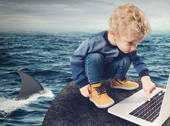 Bambino su uno scoglio circondato da squali mentre ha lo sguardo immerso nel monitor di un computer