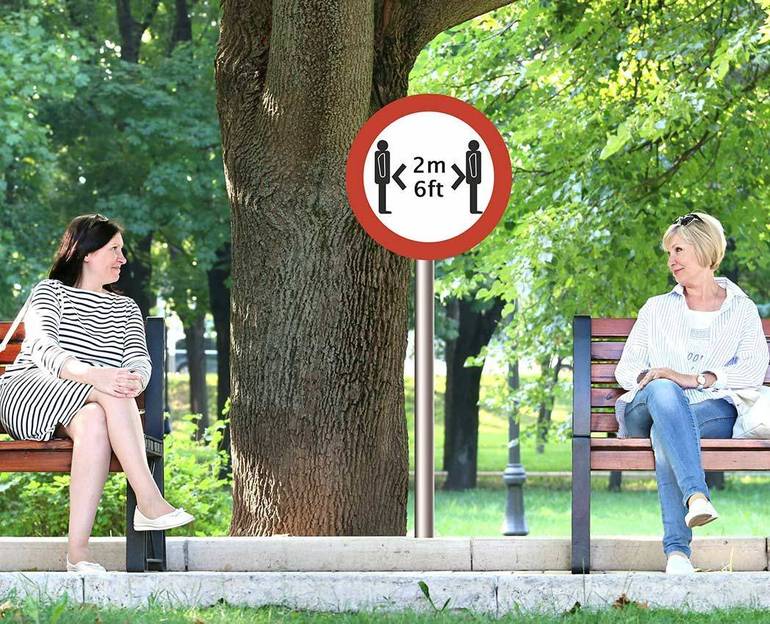 Due donne dialogano sedute su due panchine ai giardini separate da un segnale di distanziamento