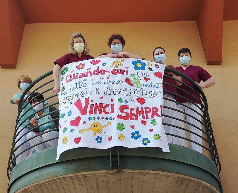Gruppo di operatori di una casa di riposo al balcone con uno striscione colorato recante la scritta: "Quando curi una malattia puoi vincere o perdere, quando ti prendi cura vinci sempre"