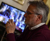 Primo piano di una persona in preghiera con il rosario, sullo sfondo uno schermo tv